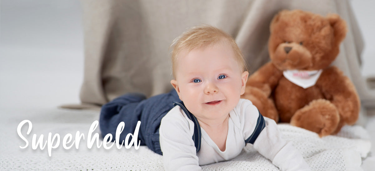 Baby mit einem Stoffbären im Hintergrund, daneben das Wort Superheld